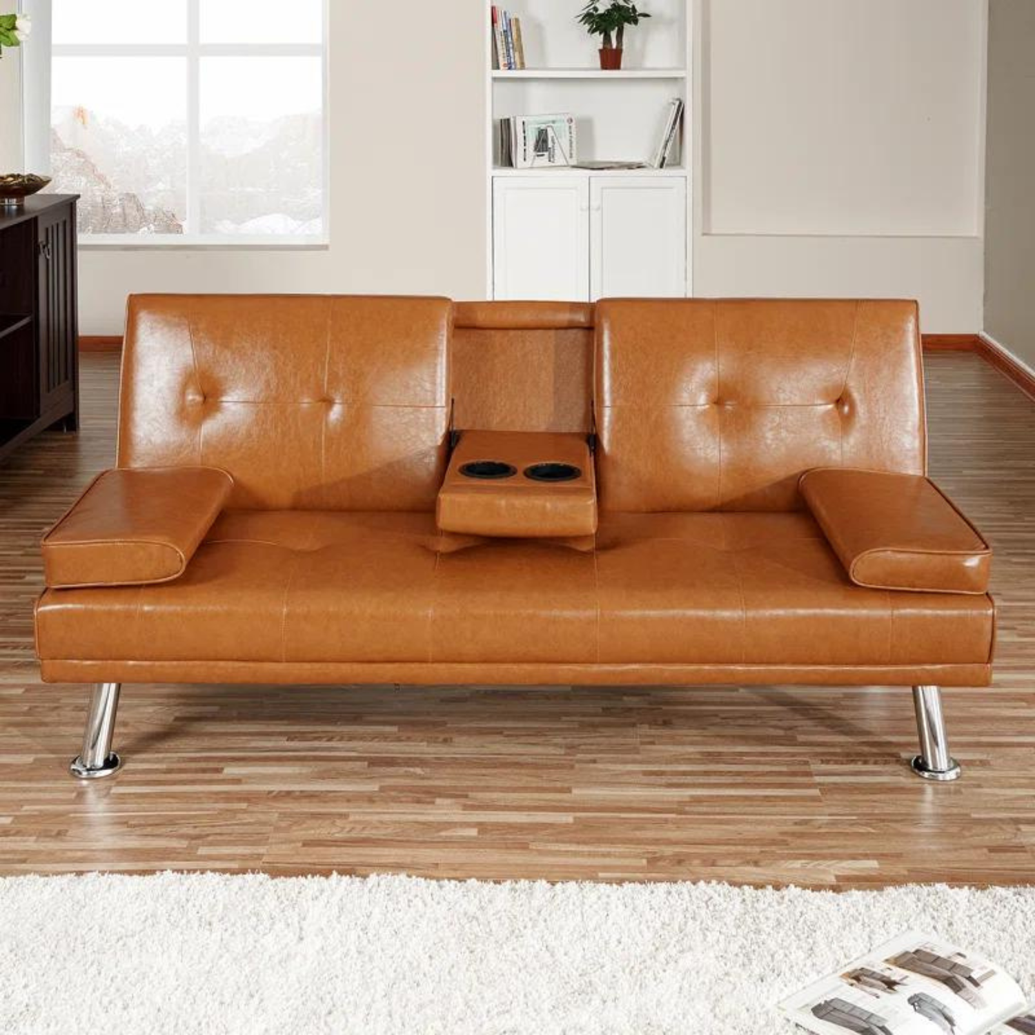 Couch Bed Futon Sleeper Sofa for Living Room | Tan Faux Leather from AHF Depot - Tan leather futon with center cupholders displayed against in a modern living room scene, offering a view out the window as well as to the bookshelf in the back.